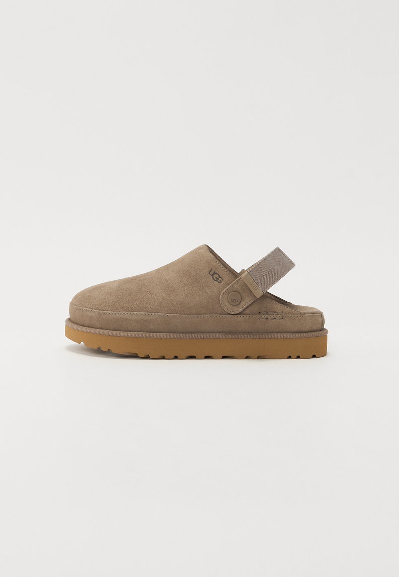 Pantof brown din suede, tip slip-on, cu talpă din cauciuc, curea pe spate și un detaliu de branding minim pe lateral. Textură netedă pe întreaga suprafață.
