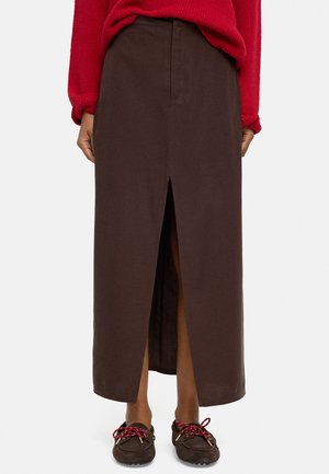 Osoba ubrana w brązową spódnicę midi z przednim rozcięciem, czerwoną bluzę i brązowe mokasyny z czerwonymi i białymi sznurowadłami, stojąca na białym tle.