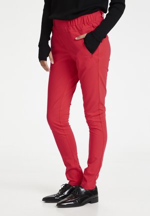 Pantalon rouge avec une taille élastique, poches latérales et coupe slim ; associé à des chaussures de ville noires. Texture de tissu lisse.