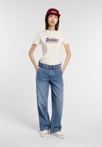 Das Model posiert in einem lässigen Outfit, trägt ein cremafarbenes Dickies-T-Shirt, blaue weit geschnittene Jeans und eine burgunderfarbene Mütze, mit den Händen in den Taschen, vor einem schlichten Hintergrund.