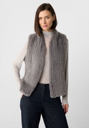 Gilet in finta pelliccia grigia con una consistenza morbida e soffice, caratterizzato da un design aperto nella parte frontale. Indossato sopra una maglia chiara e abbinato a pantaloni scuri.