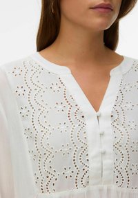 Femme portant un chemisier blanc avec une broderie œillet festonnée et trois boutons recouverts de tissu au col en V.