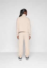 Survêtement beige en tissu doux avec un col haut, coupe décontractée et poignets côtelés. Porté avec des chaussures de sport noires.