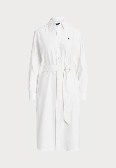 Balta medvilninė suknelė, su apykakle, ilgomis rankovėmis, mygtukų užtrauktuku ir atitinkančia juosta, skirta apibrėžti juosmenį.