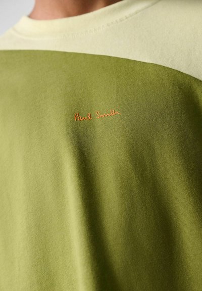 Nærbillede af en tofarvet grøn sweatshirt med lille orange broderet tekst centralt på brystet.