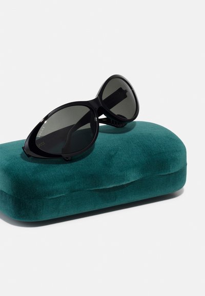 Sorte ovale solbriller som hviler på et teal fløyelsetui, med mørke linser og en slank plastramme med subtile merkevare detaljer.