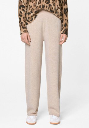 Pantalon beige à jambes larges porté avec des baskets blanches et un haut à manches longues imprimé léopard, debout devant un fond uni.