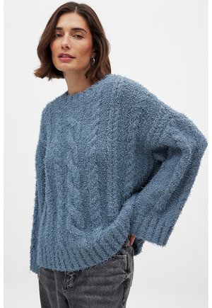 Light blue, fuzzy knit sweater with a ribbed hem and wide sleeves, featuring a textured cable pattern throughout. Paired with dark jeans.
