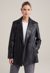 Svart läder dubbelknäppt kapp med stora skärklippta slag, sidofickor och en slät yta. Bärs över en vit topp.