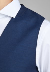 Tamno plavi prsluk od teksturirane tkanine, s oštrim ramenima i reverom, složen preko bijele košulje.