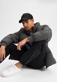 Mænds mørkegrå pufferjakke med høj krave, sort mesh baseballcap, sorte joggers og hvide sneakers med perforeret detaljering.