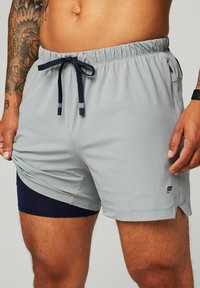 Graue Sportshorts aus leichtem Material, mit navy-blauem Kordelzug, seitlichen Schlitzen und einem internen navy-blauen Futter. Schwarzes Logo am Saum.