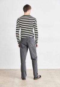 Chemise à rayures noires et blanches à manches longues, associée à un pantalon gris et des chaussures noires. Le fond est minimal avec un sol de couleur claire.