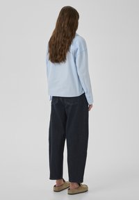 Person med långt brunt hår som bär en ljusblå skjorta, mörka lösa jeans och beigea slip-on skor, stående mot en enfärgad bakgrund.