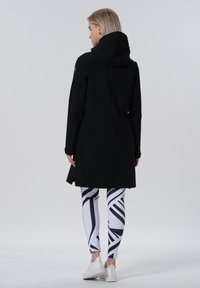 Manteau imperméable noir avec une capuche, affichant un design épuré. Assorti à des leggings à motifs blancs et noirs et des chaussures de sport blanches.