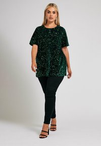 Top verde con paillettes, maniche corte, vestibilità ampia e scollatura rotonda, abbinato a pantaloni skinny neri e sandali neri con cinturini.