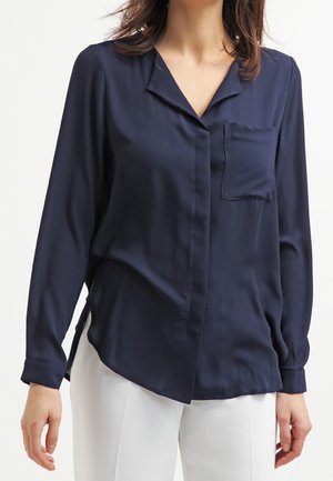 Kobieta ubrana w granatową bluzkę z długim rękawem i kieszenią z przodu, w połączeniu z białymi spodniami, na jednolitym tle.