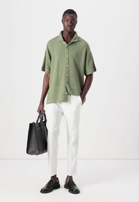 Camicia a maniche corte verde con texture, pantaloni bianchi, scarpe in pelle nera e una borsa a tracolla nera. Il modello è in piedi contro uno sfondo neutro.