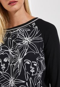 Czarny top z długim rękawem z białymi rysunkami kwiatów i linii twarzy, z żebrowaną teksturą na ramionach i okrągłym dekoltem.
