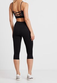 Czarny sportowy top na krótko z krzyżowanym tyłem, zestawiony z czarnymi legginsami 3/4. Modelka nosi białe trampki z szarymi akcentami.