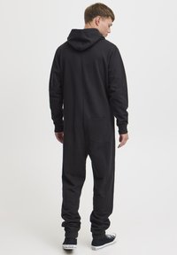 Un mono con capucha negro hecho de tejido suave, que cuenta con un bolsillo en la parte trasera y puños gather en los tobillos. Diseño sencillo y relajado.