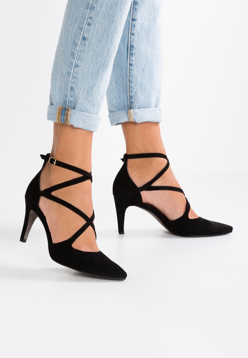 Pieds portant un jean bleu clair retroussé et des chaussures à talons hauts noires à brides sur fond blanc.