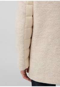 Manteau avec un corps beige à texture douce et des manches rembourrées beige clair. Le design présente une coupe ajustée et un ourlet arrondi.