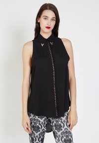 Mujer vistiendo una blusa sin mangas negra con tachuelas doradas en la parte delantera y puntas de collar decorativas, combinada con pantalones negros y blancos con estampado.