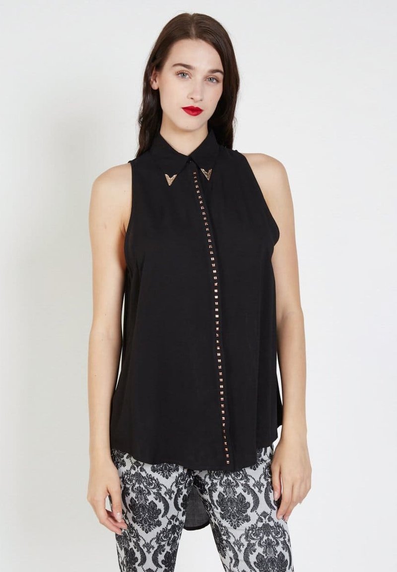 Mujer vistiendo una blusa sin mangas negra con tachuelas doradas en la parte delantera y puntas de collar decorativas, combinada con pantalones negros y blancos con estampado.