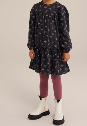Child wearing a long-sleeve black dress with pink floral print, mauve leggings, and white chunky boots standing against a plain background.