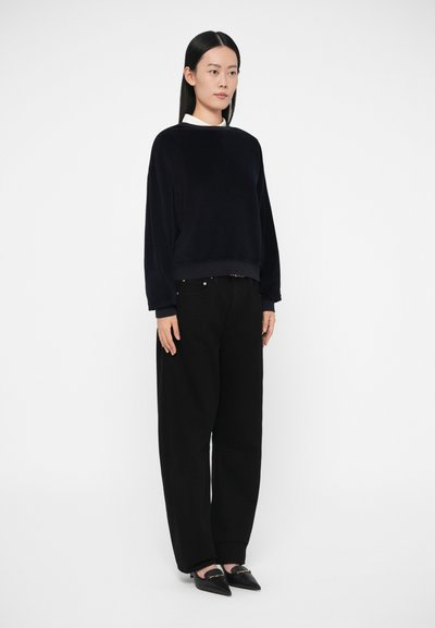 Μπλε σατέν βελούδινο φούτερ με σ�τρογγυλή λαιμόκοψη και ριμπ μανσέτες, σε συνδυασμό με μαύρο παντελόνι φαρδιάς γραμμής και μαύρα μυτερά παπούτσια.