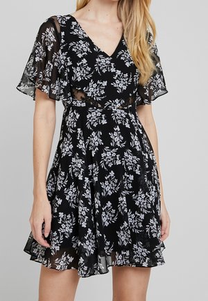 Robe noire à fleurs avec des manches transparentes, un col en V et une jupe évasée. Décorée de motifs de feuilles blanches. Tissu léger et fluide.