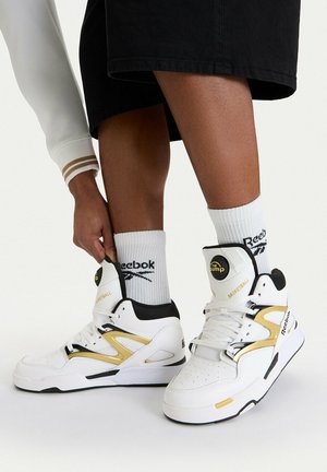Person passt weiße und goldene Reebok High-Top-Basketballschuhe an, die mit weißen Reebok Crew-Socken und schwarzen Shorts getragen werden.