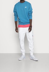 Sudadera azul claro con el logo blanco de Nike, superpuesta sobre una camiseta rosa, combinada con joggers blancos con bolsillos laterales y zapatos a juego.
