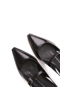 Zapatos Mary Jane de charol negro con punta afilada, que cuentan con una delgada correa en el tobillo y una hebilla plateada. Acabado liso y brillante.