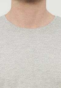 Λαιμός και άνω μέρος του στήθους ενός ατόμου που φοράει ανοιχτό γκρι T-shirt με στρογγυλή λαιμόκοψη, πάνω σε απλό φόντο.