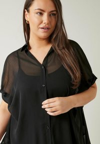 Kobieta z długimi brązowymi włosami, nosząca złote kolczyki w kształcie kół i przezroczystą czarną bluzkę na guzikach na czarnym topie, na jednolitym tle.