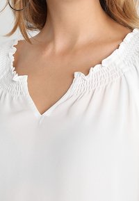 Kvinna som bär blus i vitt med samlad, volangkrage och V-formad skärning, som visar en del av halsen och axeln med en synlig hoop örhänge.