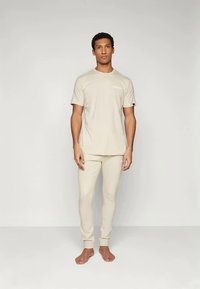 Bež pamučna majica s kratkim rukavima i malim logotipom, u kombinaciji s uskim trenirkama koje imaju rebraste manžete i glatku teksturu.