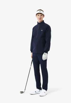 Marineblauw golftrainingspak met een jas met ritssluiting en lange broek, aangevuld met een witte zonneklep, witte golfhandschoenen en witte sneakers.