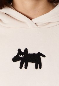 Felpa color crema con applicazione di un cane nero testurizzato sul davanti, caratterizzato da orecchie prominenti e una coda, che aggiunge un interessante elemento visivo.