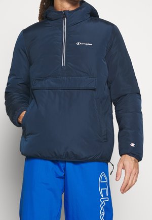 Mann trägt eine marineblaue gesteppten Kapuzenjacke von Champion und leuchtend blaue Champion-Shorts mit Logo am Bein und am Handgelenk.