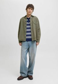 Un jeune homme portant une chemise oversize vert olive, un pull rayé bleu marine, un jean clair à jambes larges et des chaussures marron foncé, se tenant contre un fond blanc.
