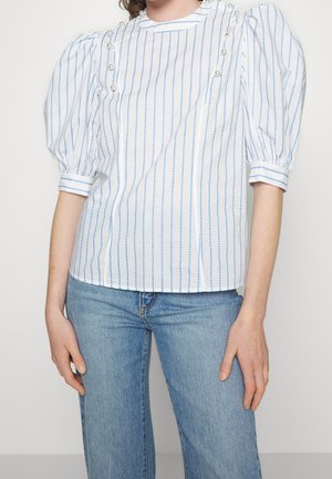 Blouse blanche à rayures verticales bleues, manches bouffantes mi-longues aux coudes, et boutons en perle près des épaules, portée avec un jean bleu.