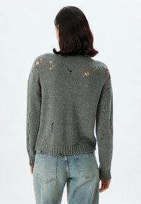 Grijze, gebreide trui met een geribde zoom en manchetten, met overal versleten gaten. Gecombineerd met lichtgewassen denim jeans.