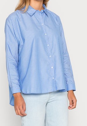 Woman wearing a loose-fitting light blue shirt with white vertical stripes and long sleeves, paired with light blue jeans.