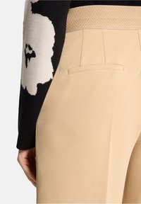 Pantalon beige à texture lisse, doté d'une poche arrière unique et d'une ceinture à motif chevron, associé à un pull noir.