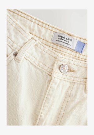 Wide leg, high-rise jeans made of soft, off-white fabric. Features stitched seams and a metal button with brand detailing and label.