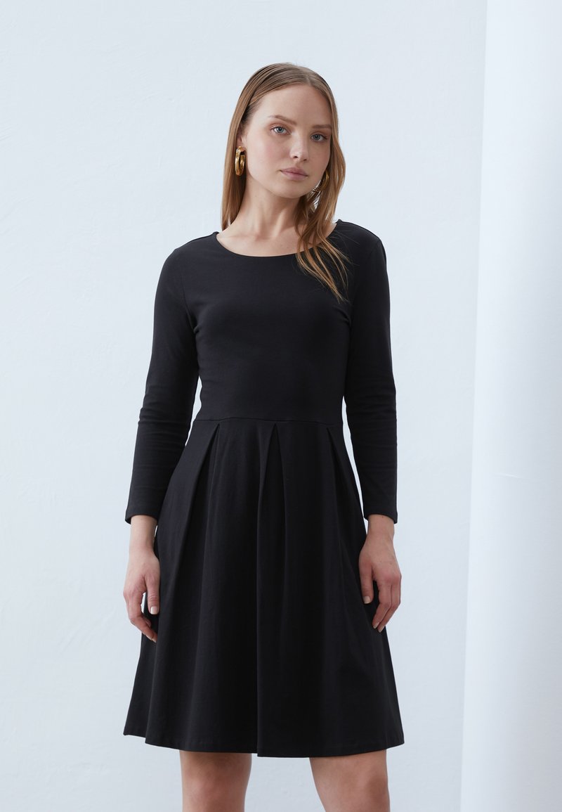 Black long-sleeve dress with a fitted bodice and flared, pleated skirt. Smooth cotton fabric; classic round neckline and simple design.