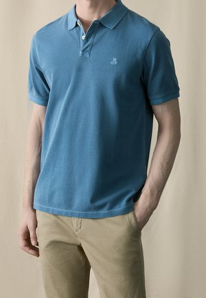 Homme portant un polo bleu à manches courtes avec un petit logo et un pantalon beige, debout avec une main dans la poche.
